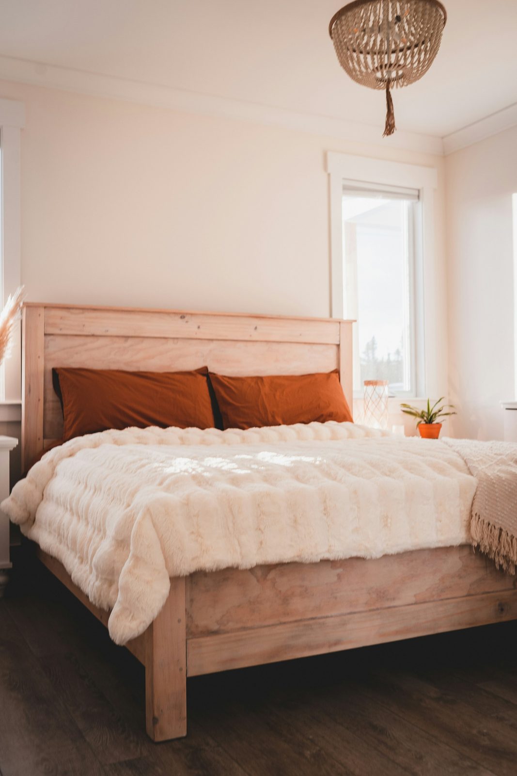 A double bed with a pale wooden headboard and rust-coloured cushions, a woven pendant light and a tall window in a white room