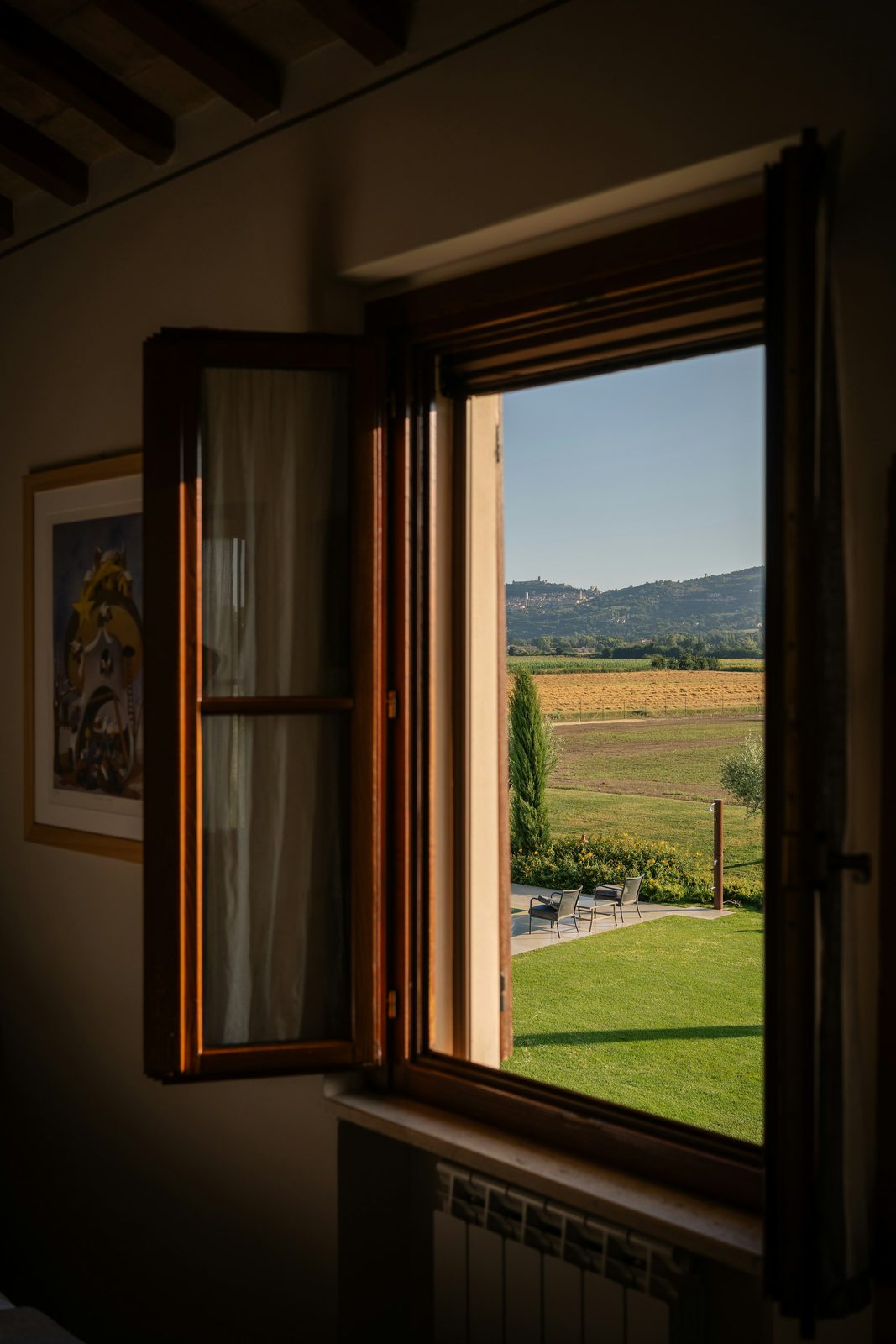 A tall shuttered window open onto a lawn and a green hill, in a room with a dark ceiling beam