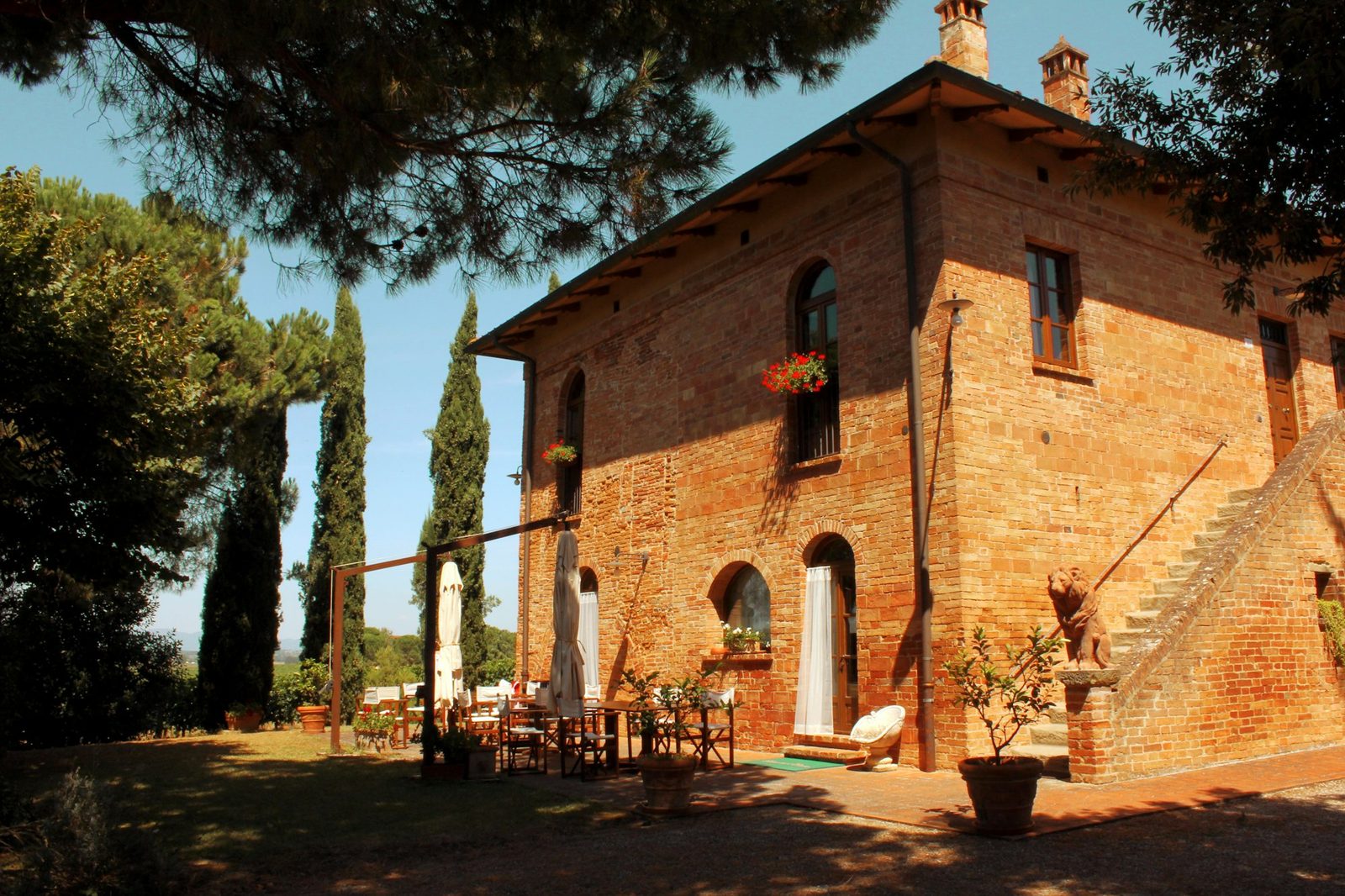The villa's warm brick and stone front with an outside staircase, tall cypresses and umbrella pines, and a table set in the shade