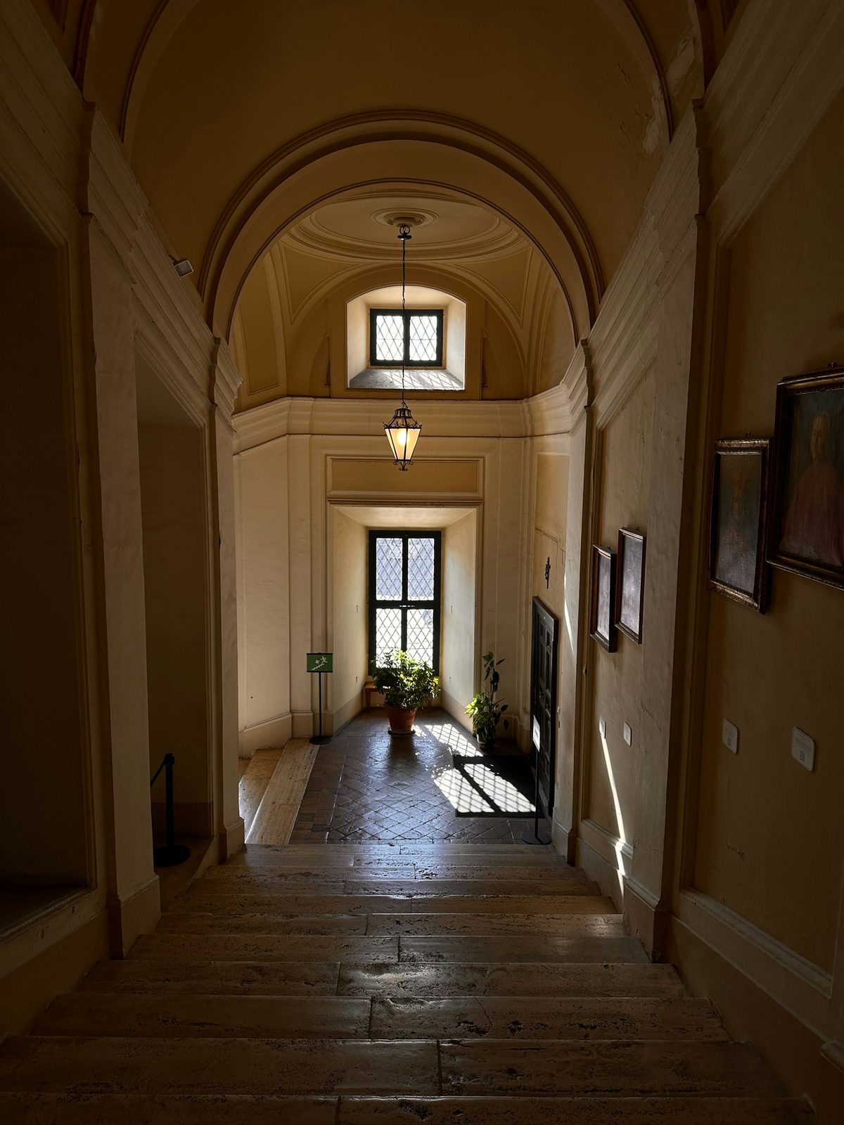 A vaulted corridor with a stone floor, arched doorways and a lantern, ending in a small window