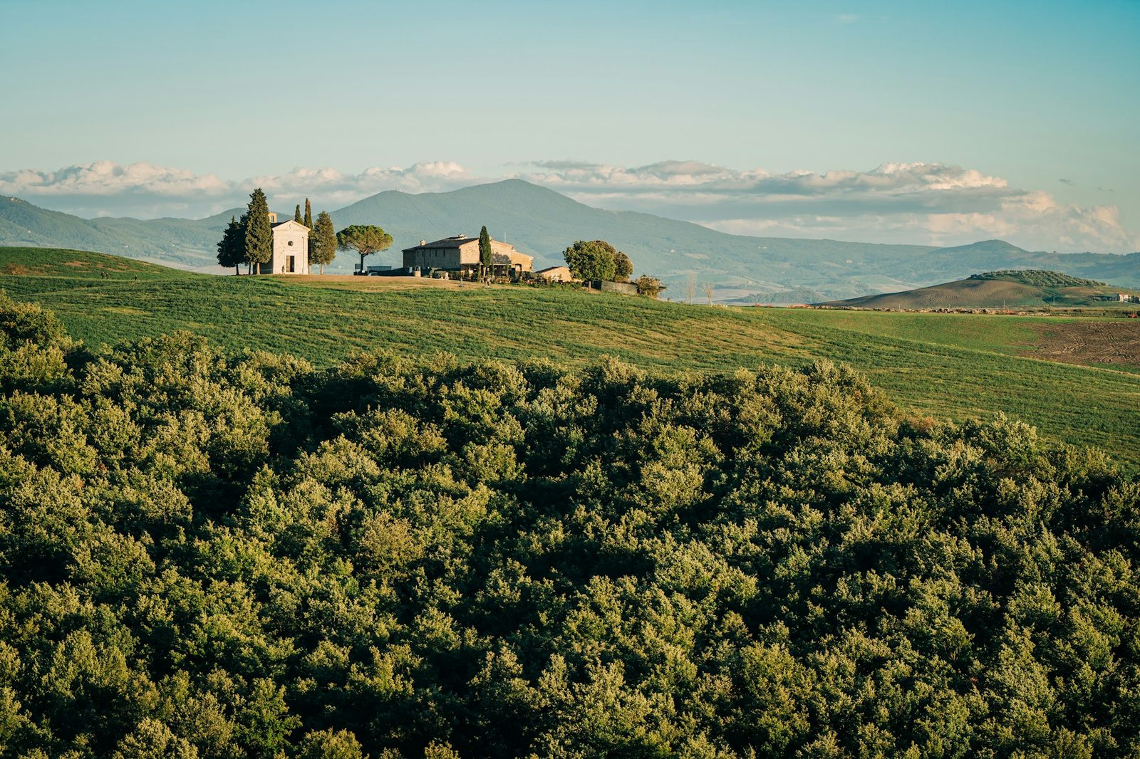 A small chapel and farmhouse on a green hill with a wooded ridge behind