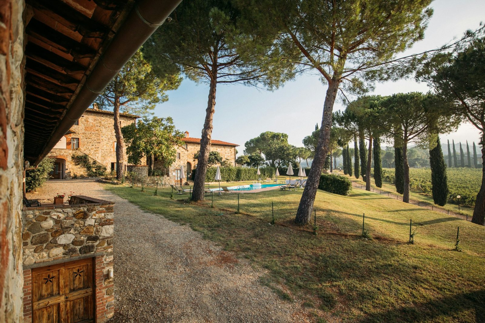 A gravel track beside a stone outbuilding under umbrella pines, with a row of cypresses, a lawn and the pool in the distance