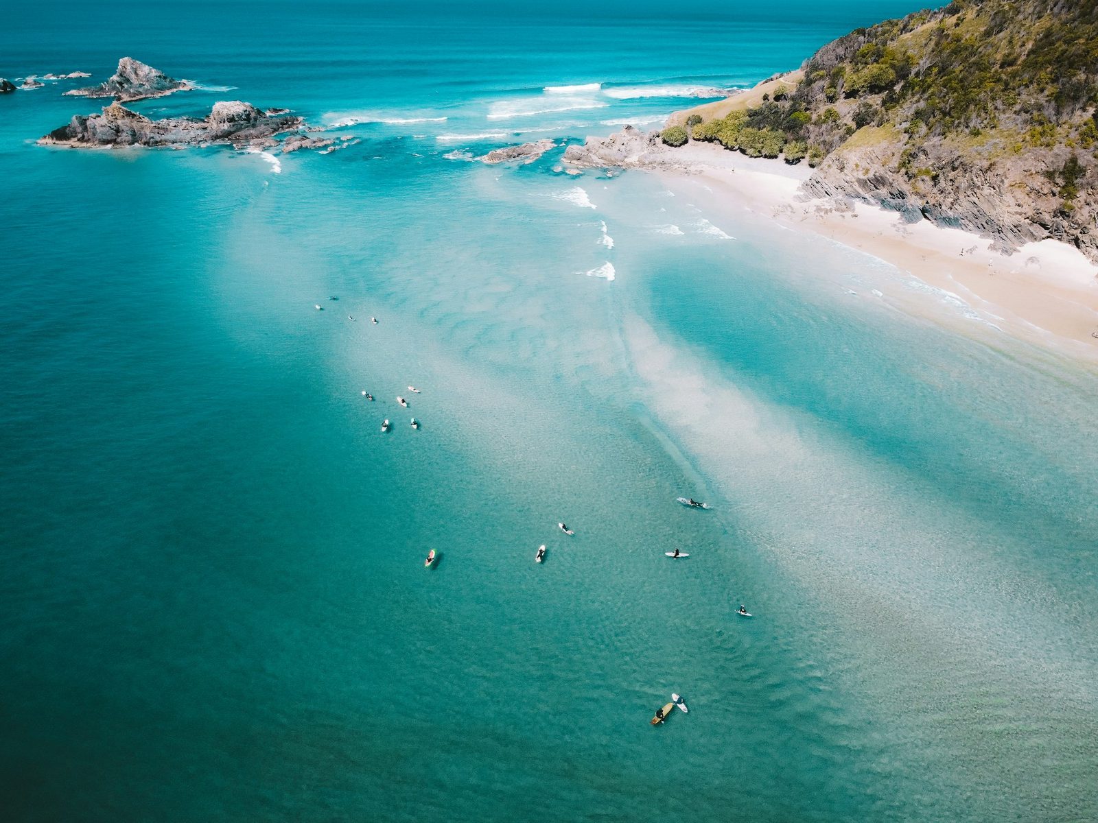 Aerial view of surfers in pale turquoise water beside a rocky headland.