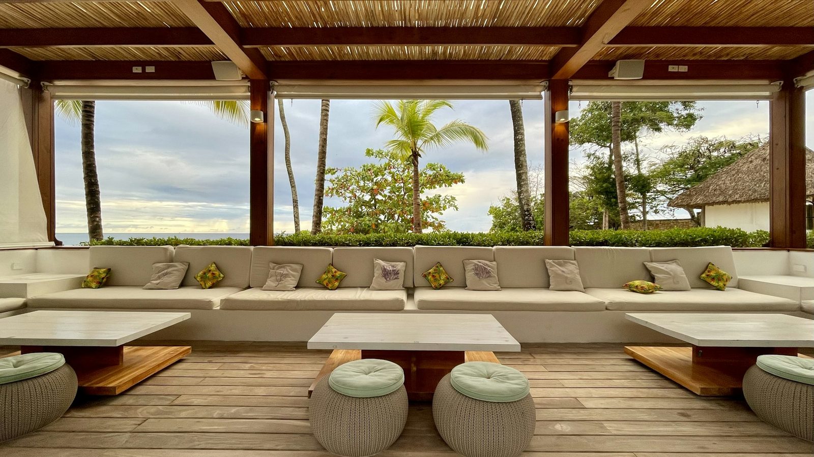 A covered terrace with a long cushioned sofa and round woven stools, open to palms.