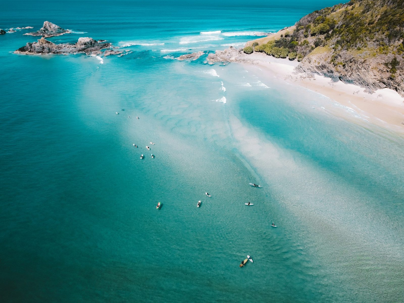 Aerial view of surfers in pale turquoise water beside a rocky headland.