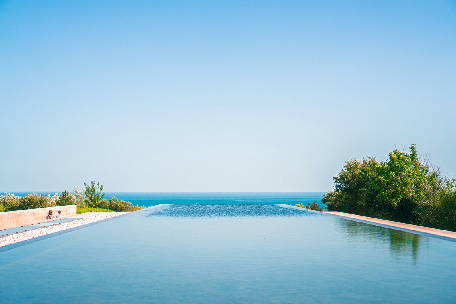 A long infinity pool whose far edge meets the horizon, with green shrubs to one side.