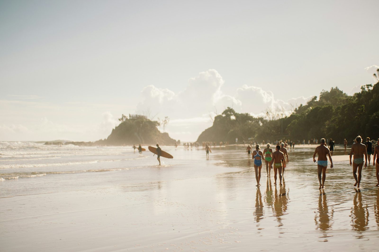 People walking along a wet beach in hazy light, with a surfer and a headland behind.