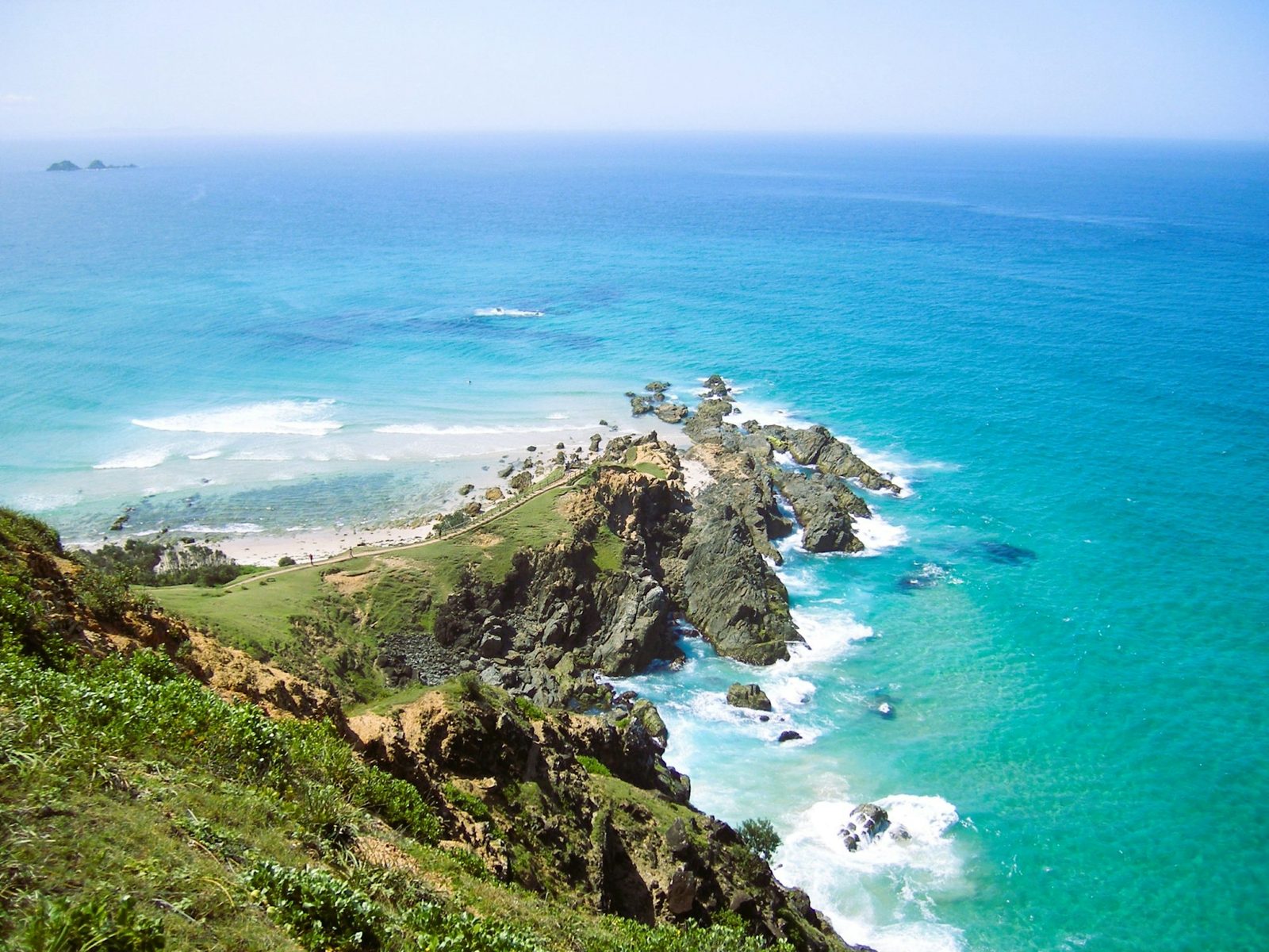 A grassy clifftop above a rocky point and a bright turquoise sea, with an island on the horizon.
