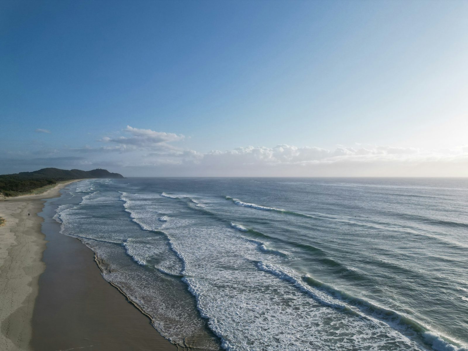 An aerial view of long lines of waves breaking on a beach under a pale blue sky.