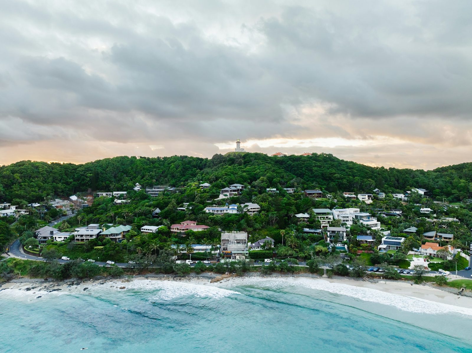 An aerial view of a green headland with houses on its slopes, a white-sand beach in front and a small lighthouse on the skyline.