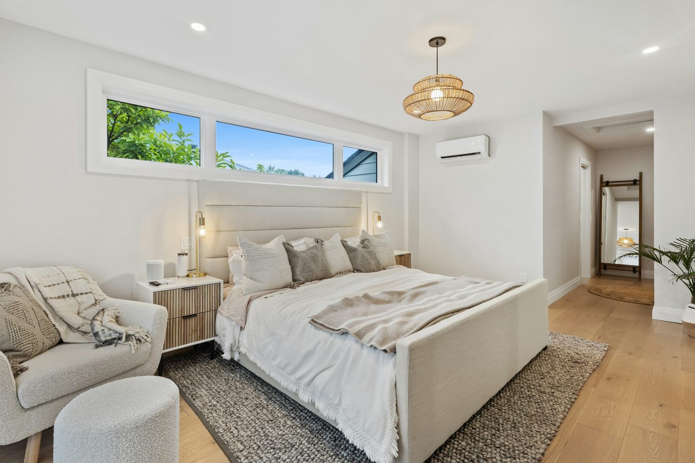 A bedroom with a queen bed in pale linen, an armchair and stool, a woven pendant light and a long window above the bed