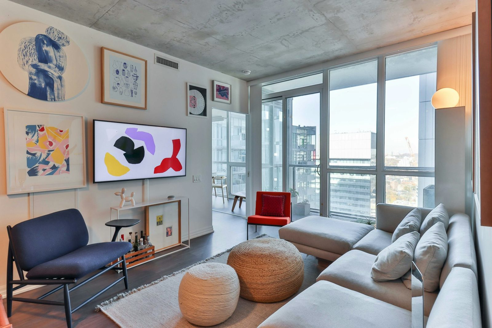 A living room with floor-to-ceiling windows, a grey sectional sofa, a blue armchair and colourful framed prints, with city buildings outside