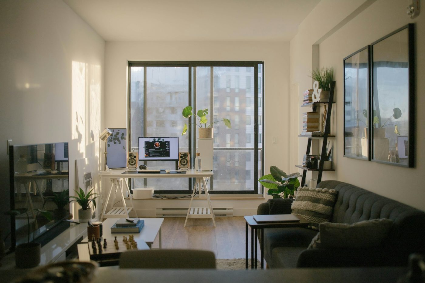 A desk with a monitor and speakers beside a large window, with a grey sofa and plants in the room
