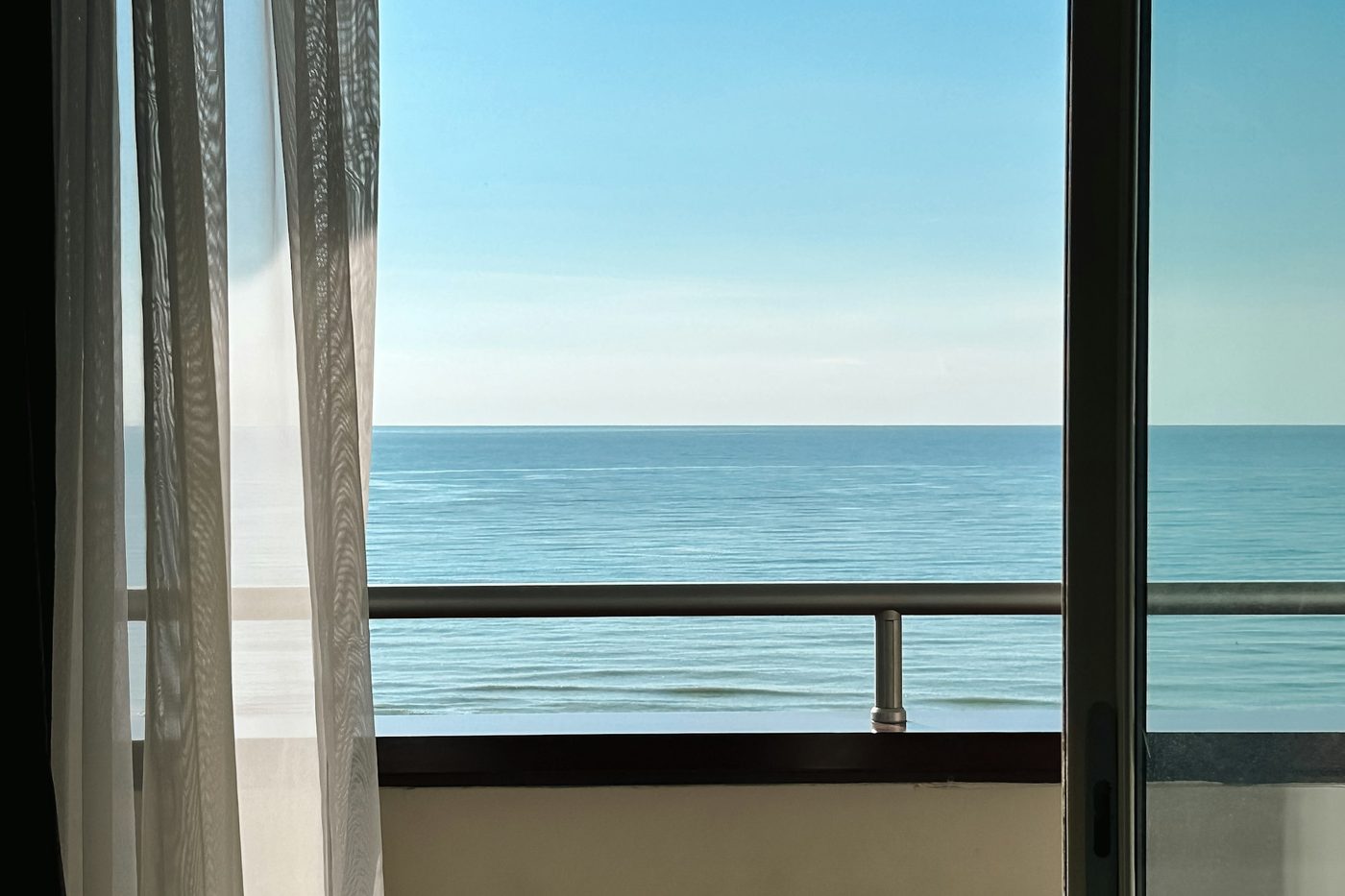 A sheer curtain and an open glass door onto a balcony railing, with the sea and a blue sky beyond