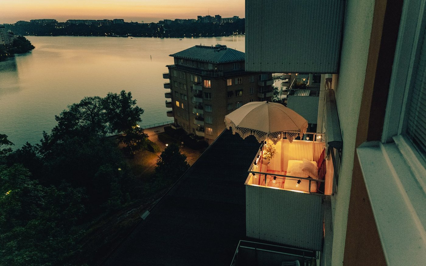 A lit balcony with seating under a fringed parasol at dusk, looking over calm water and trees