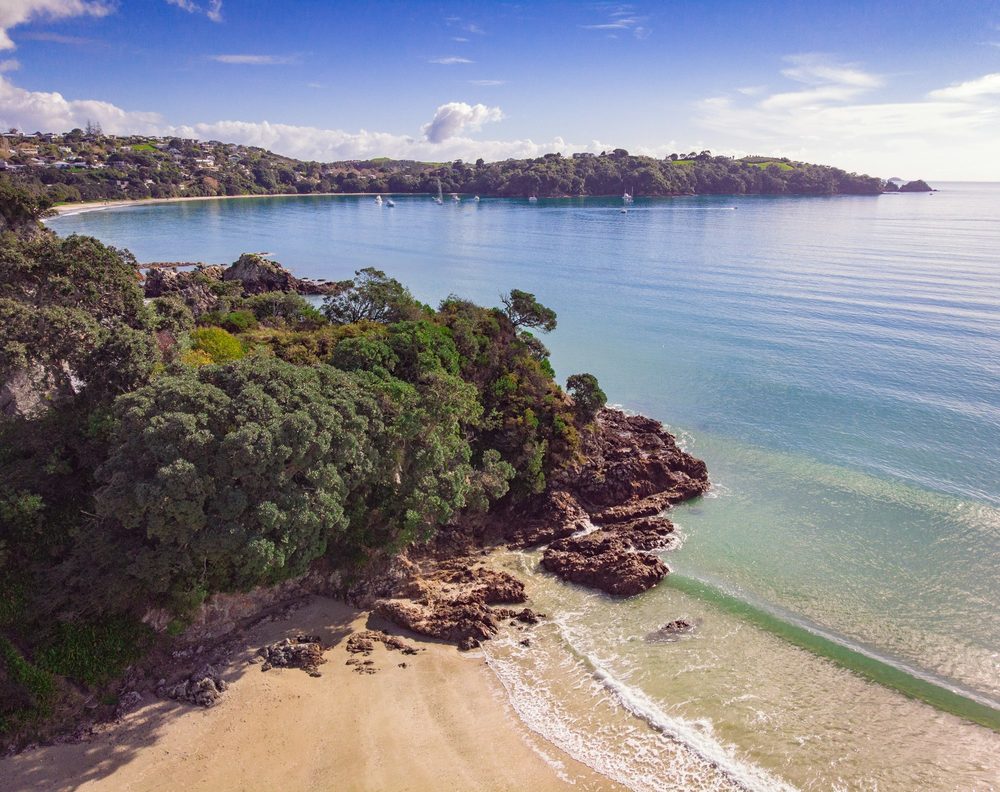 A bay of bush, rock and pale sand beside calm water, seen from above